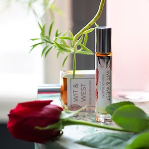 May include: A clear glass roll-on perfume bottle with a silver cap and a label that reads "WIT & WEST ROSA DE BOLERO". A red rose and green plant are in the foreground. Another perfume bottle is in the background.