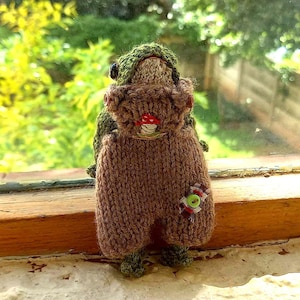 May include: A knitted gnome doll with a green hat and brown dungarees. The gnome features a red mushroom button and a small decorative button. The doll is positioned on a windowsill, with a blurred green backdrop.