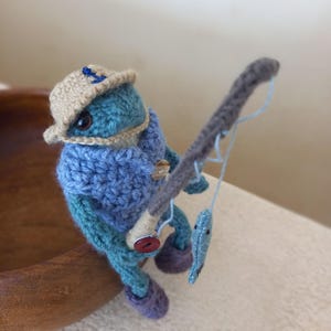 Op de afbeelding: Een gehaakte kikker met een bruine hoed met een blauwe anker, een blauwe trui en blauwe laarzen houdt een hengel vast met een blauwe vis aan de lijn.