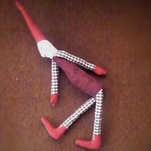 May include: A red and white fabric doll with a long nose and a burgundy body. The doll is wearing a black and white checkered pattern on its legs and arms.