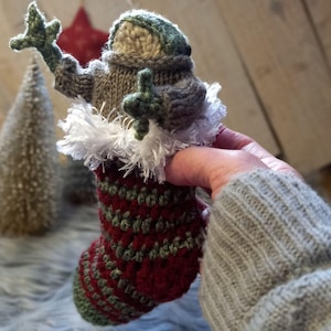 May include: A crocheted gnome wearing a gray and green hat and holding a green leaf. The gnome is sitting in a red and green striped stocking with a white fur trim.