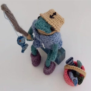 May include: A crocheted frog wearing a blue sweater and a tan hat with an anchor on it. The frog is holding a fishing rod with a blue fish on the line. The frog is sitting on a blue box and there is a pink bucket with blue and green fish inside.