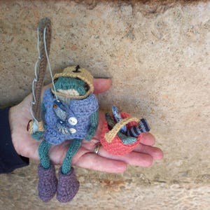 May include: A crocheted frog wearing a brown and yellow hat holds a fishing rod and a red basket filled with crocheted fish.