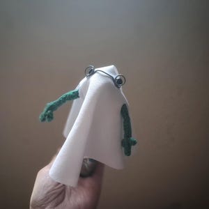 May include: A whimsical handmade frog toy, with green crocheted arms and a white fabric body. The frog has wire-framed eyes and is held in a hand. The toy is a unique, handcrafted item.