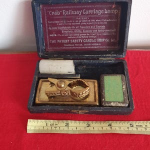 May include: An open, antique blue box containing a brass candle holder, a white candle, and a green rectangular object. The box has text that reads "Crab Railway Carriage Lamp" and "Patent". A ruler shows measurements in inches.