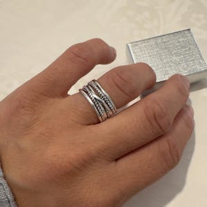 Op de afbeelding: Een brede zilveren ring met een ontwerp van meerdere banden, bestaande uit twee gladde buitenbanden en twee centrale gedraaide touwstructuur-banden die elkaar kruisen. De ring heeft een gepolijste metalen afwerking en een kleine geschenkdoos.