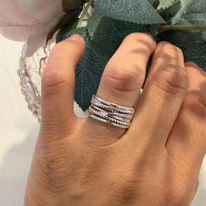 Op de afbeelding: Een brede zilveren ring met een ontwerp van meerdere banden met afwisselende texturen. De ring bestaat uit verschillende gestapelde banden, waaronder glad gepolijst zilver, een kralentextuur en een gedraaid touwpatroon, wat een gelaagd uiterlijk creëert.