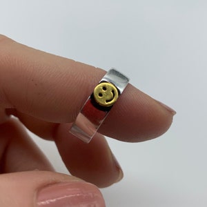 May include: A silver ring with a gold smiley face charm. The ring is worn on a finger.