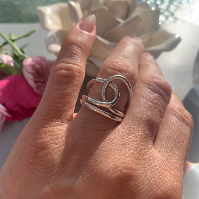 Op de afbeelding: Een zilveren ring met een geknoopt ontwerp. De ring wordt aan een vinger gedragen.