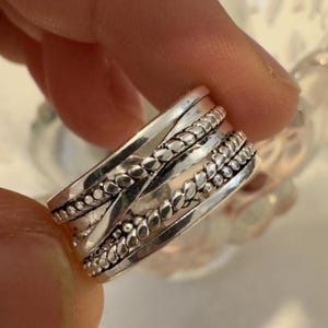 Pode incluir: Um anel largo de tom prateado com um design de múltiplas bandas, apresentando duas bandas trançadas tipo corda que se cruzam sobre uma banda central de contas. O metal tem um acabamento polido com áreas rebaixadas escuras.