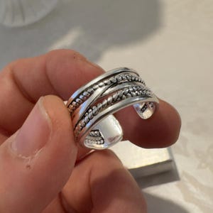 May include: A silver-coloured open band ring featuring a multi-layered design with two twisted rope-style bands crossing over a central beaded band. The metal surface has a polished finish, and the interior of the band is stamped with 925.