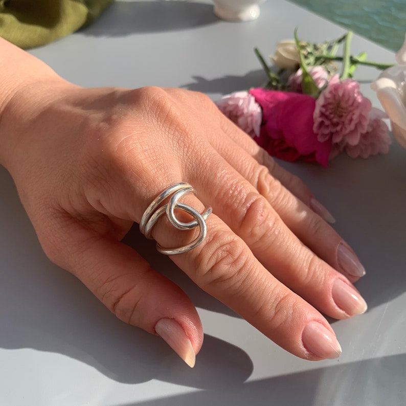 Op de afbeelding: Een zilveren ring met een knoopontwerp. De ring wordt aan een vinger gedragen.