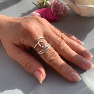 Op de afbeelding: Een zilveren ring met een uniek, verweven ontwerp. De ring wordt gedragen op een vinger met lichtroze nagellak.