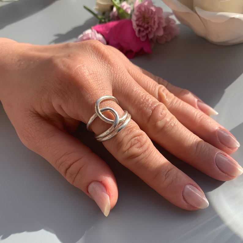 Op de afbeelding: Een zilveren ring met een uniek, verweven ontwerp. De ring wordt gedragen op een vinger met lichtroze nagellak.