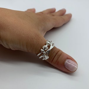 May include: A silver ring with a unique interwoven design. The ring is worn on a person's thumb.