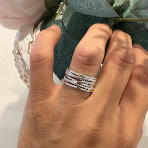May include: A wide silver ring featuring a multi-band design with alternating textures. The ring consists of several stacked bands, including smooth polished silver, a beaded texture, and a twisted rope pattern, creating a layered appearance.