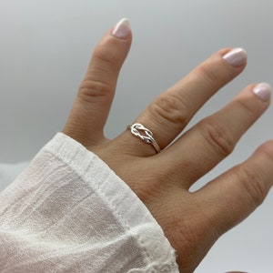 Op de afbeelding: Een zilveren ring met een geknoopt ontwerp. De ring wordt aan een vinger gedragen.