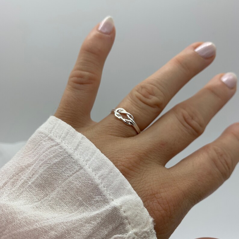 Op de afbeelding: Een zilveren ring met een geknoopt ontwerp. De ring wordt aan een vinger gedragen.
