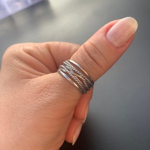 May include: A silver ring with a braided design. The ring is worn on a person's thumb.