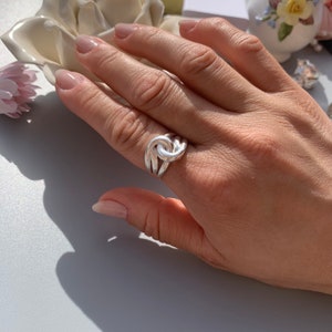 May include: A silver ring with a knot design. The ring is worn on a person's finger.