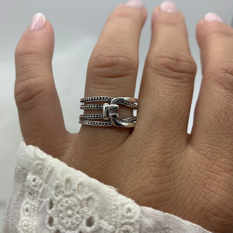 May include: A silver ring with a multi-layered design featuring a buckle detail. The ring is adorned with small beads.