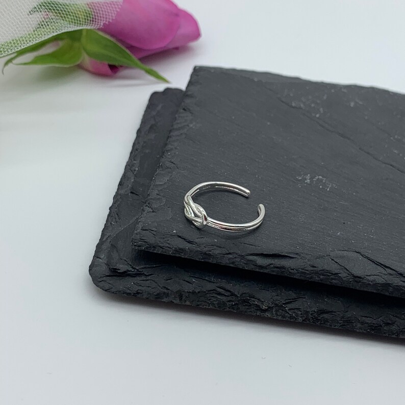 Op de afbeelding: Een zilveren teenring met een knoopdesign. De ring ligt op een zwart leisteen oppervlak.