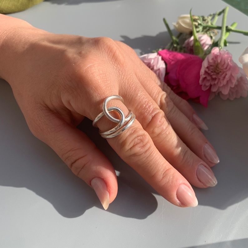Op de afbeelding: Een zilveren ring met een dubbel lusontwerp. De ring wordt gedragen op een vinger met lichtroze nagellak.