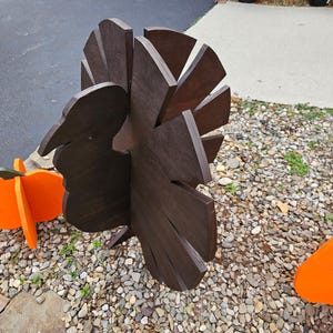 May include: A dark brown wooden turkey decoration with a fan-shaped tail and a carved head. Beside it are orange pumpkin decorations with green stems. The decorations are placed on a bed of small stones.