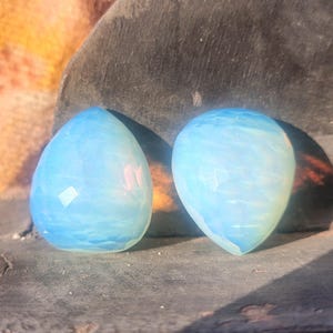 May include: Two opalescent, teardrop-shaped gemstones with a light blue hue. The stones have a faceted surface, catching the light and creating a shimmering effect. The gems are set against a dark, textured background.