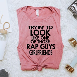 May include: A pink sleeveless tank top with the text "Tryin' to look like one of those rap guys girlfriends" printed in black.