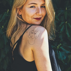 Pu&ograve; includere: Una donna con lunghi capelli biondi ha un tatuaggio di un cane in stile lineare sulla spalla.