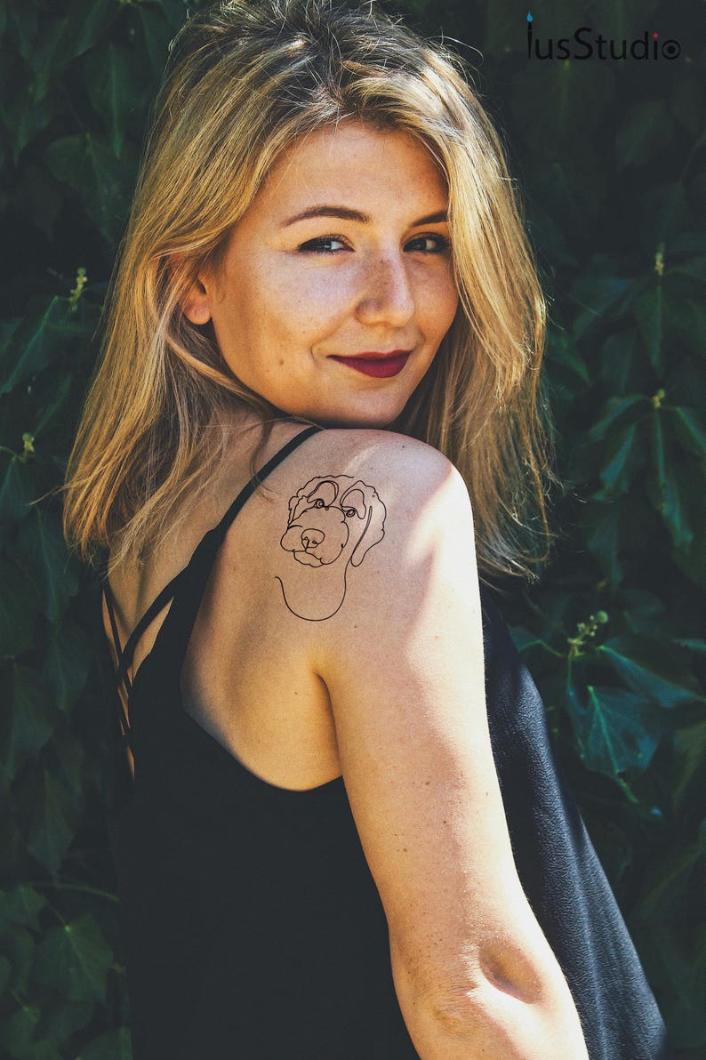 Pu&ograve; includere: Una donna con lunghi capelli biondi ha un tatuaggio di un cane in stile lineare sulla spalla.