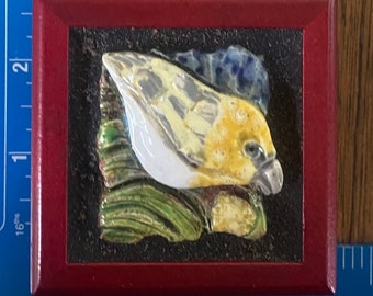 May include: A ceramic tile featuring a yellow and white bird in a green and brown leafy setting. The tile is framed in a dark red wooden frame.