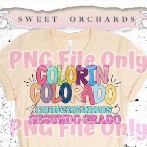 May include: Cream-colored t-shirt with the words "COLORIN COLORADO" in colorful letters. Below the text are the words "comenzamos SEGUNDO GRADO" in pink and teal. The shirt has rolled-up sleeves and a crew neck.