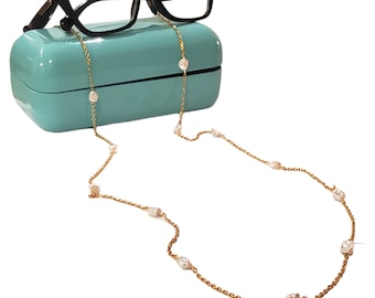 May include: Black eyeglasses rest on a teal case, with a gold chain eyeglass holder draped below. The chain features small, irregularly shaped, off-white stones spaced along its length. The glasses have a thick, black frame.