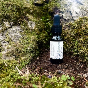 May include: A small, dark brown glass bottle with a black dropper top. The bottle is labeled with white text and a black and white illustration of a plant. The bottle is sitting on a bed of brown dirt and green moss.