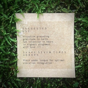 Op de afbeelding: Een bruine papieren kaart met instructies voor het gebruik van een product. De tekst luidt: "SUGGESTED USE Visualize grounding gratitude in Earth Set intention in heart in highest alignment with love ///. SHAKE SEVEN TIMES 3DROPS Place under tongue for optimal vibration integration"