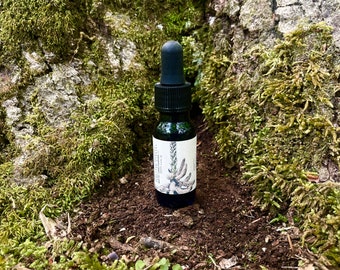 Op de afbeelding: Een kleine, donkere glazen fles met een zwarte druppeltop. De fles heeft een wit etiket met botanische illustraties. De achtergrond toont een met mos bedekte boomstam en donkere aarde.