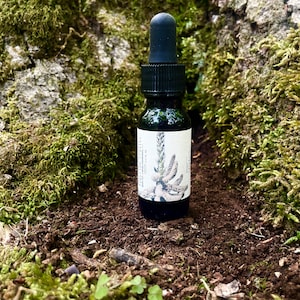 Könnte beinhalten: Eine kleine, dunkelbraune Glasflasche mit schwarzem Tropfer. Die Flasche hat ein weißes Etikett mit einer Schwarzweißillustration einer Pflanze. Die Flasche steht auf einem Bett aus brauner Erde und grünem Moos.