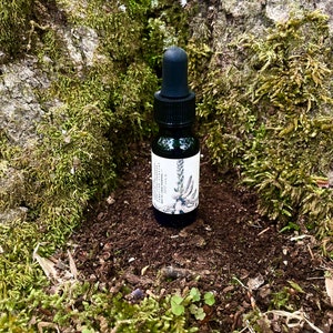 Puede incluir: Una pequeña botella de vidrio marrón oscuro con un gotero negro y una etiqueta blanca. La etiqueta tiene una ilustración en blanco y negro de una planta y el texto "Wildcrafted Botanicals". La botella está sentada en un lecho de tierra marrón y musgo verde.