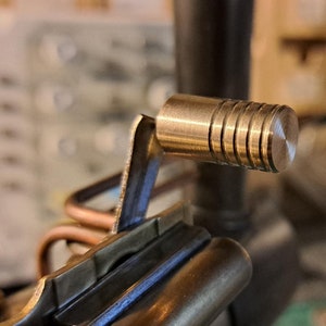 Könnte beinhalten: Nahaufnahme eines Messinghebels mit einem gerändelten Knopf. Der Hebel ist an einem größeren Messingobjekt befestigt.