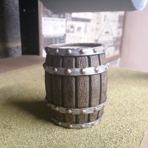 Puede incluir: Un barril de madera en miniatura con bandas y remaches metálicos. El barril es marrón con vetas de madera verticales y bandas plateadas. Los remaches de color cobre aseguran las bandas. El barril está sobre una superficie verde.