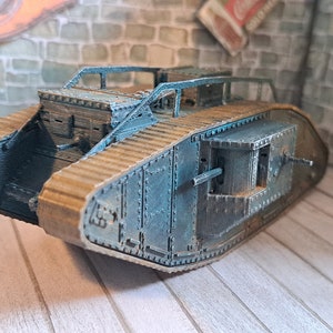 Op de afbeelding: Een kartonnen model van een Britse Mark IV tank uit de Eerste Wereldoorlog, geschilderd in een blauwgrijze kleur. De tank staat op een houten oppervlak.
