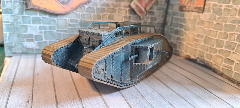 Op de afbeelding: Een kartonnen model van een Britse Mark IV tank uit de Eerste Wereldoorlog, geschilderd in een blauwgrijze kleur. De tank staat op een houten oppervlak.