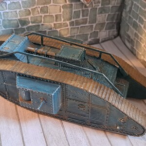 Op de afbeelding: Een blauw en bruin model van een tank uit de Eerste Wereldoorlog met een gedetailleerd ontwerp. De tank heeft een verhoogd platform met een geschutskoepel en een ladder aan de zijkant. De tank staat op een houten oppervlak.