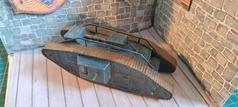 Op de afbeelding: Een blauw en bruin model van een tank uit de Eerste Wereldoorlog met een gedetailleerd ontwerp. De tank heeft een verhoogd platform met een geschutskoepel en een ladder aan de zijkant. De tank staat op een houten oppervlak.