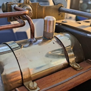 Könnte beinhalten: Nahaufnahme eines Messing-Dampfkessels mit einem Kupferrohr und einem Messingventil. Der Kessel ist auf einem Holzrahmen montiert.