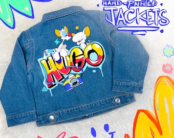 May include: A blue denim jacket with a hand-painted design. The back of the jacket features cartoon characters, the name "HUGO" in graffiti style, and a camera graphic. The text "CUSTOM HAND PAINTED JACKETS" is above the design.