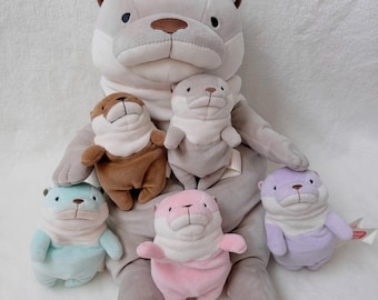 May include: A large gray plush otter with six smaller otter plush toys in various colors: brown, gray, blue, pink, and purple. Each otter has a white scarf and a cute, friendly expression. The plush toys are soft and cuddly.