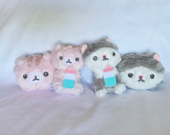 May include: Three plush toys, each with a different color and design. The first toy is pink with a brown nose and eyes, the second is white with a pink nose and eyes, and the third is gray with a white nose and eyes. Each toy is holding a small, blue and pink bottle.